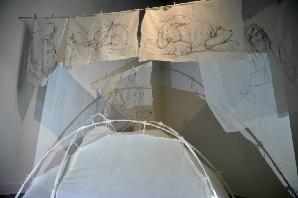 This photo shows Tatiana Garmendia’s exhibit, “No Hiding Place Down Here”. The centerpiece is a skeletal frame of a tent with thin covering, and hung above are scrim depicting art of poverty.